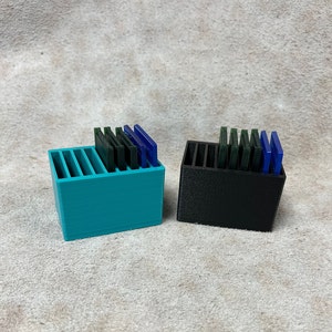 May include: Two small, rectangular storage containers. One is teal, the other black. Each has vertical dividers and holds several rectangular, colored objects in shades of green and blue. The containers sit on a textured, light-colored surface.