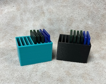May include: Two small, rectangular storage containers. One is teal, the other black. Each has vertical dividers and holds several rectangular, colored objects in shades of green and blue. The containers sit on a textured, light-colored surface.