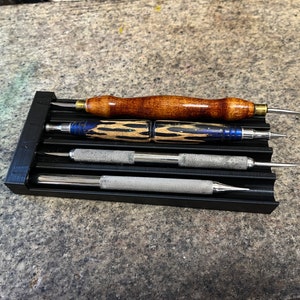 May include: A black organizer tray holding four leatherworking tools. The tools include a wooden handle with a brass ferrule, a blue and wood grain handle, and two silver metal handles. The tools have sharp, pointed tips.