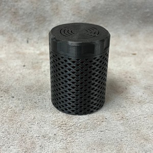 Op de afbeelding: Een zwarte cilindrische container met een roosterontwerp op de behuizing en een cirkelvormig patroon op het deksel. De container is gemaakt van een mat materiaal en staat op een getextureerd oppervlak.