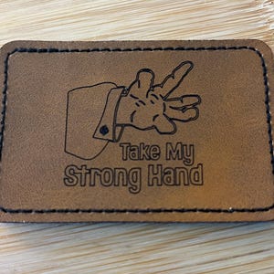 May include: A rectangular brown leather patch with black stitching around the edges. The patch features a drawing of a hand with a suit sleeve and the text "Take My Strong Hand" in a bold font.