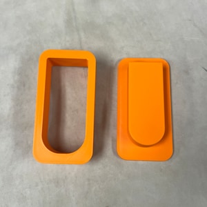 May include: Two bright orange, 3D-printed objects. One is a rectangular frame, and the other is a rectangular shape with a curved top and a clip-like design. The objects are likely for crafting or DIY projects.