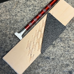 May include: A close-up of leather crafting tools and materials. A red metal mallet and a beige leather strip with a geometric pattern are visible. A black, triangular piece of abrasive material is also present, suggesting leatherworking.
