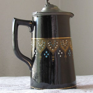 May include: A black ceramic pitcher with a silver lid and a gold and blue geometric design. The pitcher has a handle and a spout.
