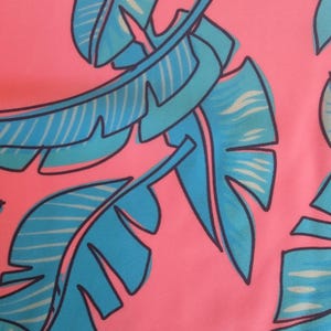 Könnte beinhalten: Nahaufnahme eines pinkfarbenen Stoffes mit einem sich wiederholenden Muster aus blau und hellblau stilisierten tropischen Blättern. Die Blätter haben dunkelblaue Umrisse und weiß gestreifte Details, die ein lebendiges, sommerliches Design erzeugen.