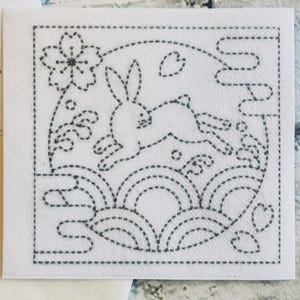 Könnte beinhalten: Eine Schwarzweiß-Zeichnung eines Kaninchens, das über einen Mond mit einer Blume und Wellen im Hintergrund springt. Das Design ist auf einem weißen Stoffhintergrund.