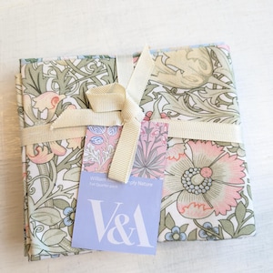 May include: A bundle of patterned fabric fat quarters tied with a cream ribbon. The fabric displays a floral design in pink, blue, and green on a white backdrop. A tag with the V&A logo and text is attached to the bundle.