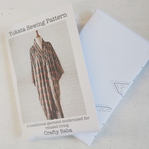 May include: A sewing pattern for a Yukata, a traditional Japanese garment, with a printed image of a long, loose-fitting robe with a colourful pattern. The text on the pattern reads "Yukata Sewing Pattern" and "A traditional garment modernised for relaxed living Crafty Baba".