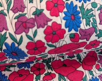 May include: Close-up of a floral fabric featuring a repeating pattern of red poppies, blue daisies, and purple bellflowers on a cream background. Green leaves and stems intertwine among the flowers, creating a vibrant and colorful design.
