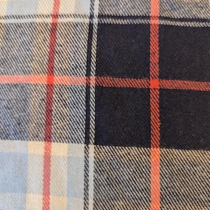 May include: Close-up of a plaid fabric with a pattern of intersecting lines. The design features a combination of light blue, navy blue, white, and red colors. The fabric has a textured appearance, suggesting a woven material.