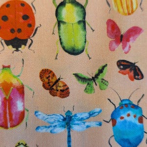 May include: Peach-coloured fabric with a repeating pattern of insects. The design includes ladybirds, beetles, butterflies, and dragonflies in red, green, blue, orange, and yellow. The insects are painted in a watercolour style.