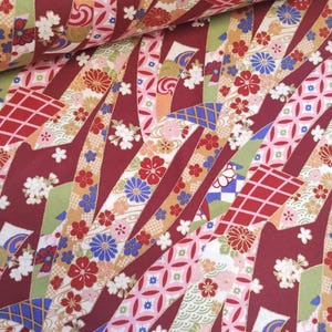 May include: Red fabric with a repeating pattern of colourful ribbons and flowers. The ribbons are arranged in a diagonal pattern and are decorated with gold accents. The flowers are in shades of pink, blue, and white.