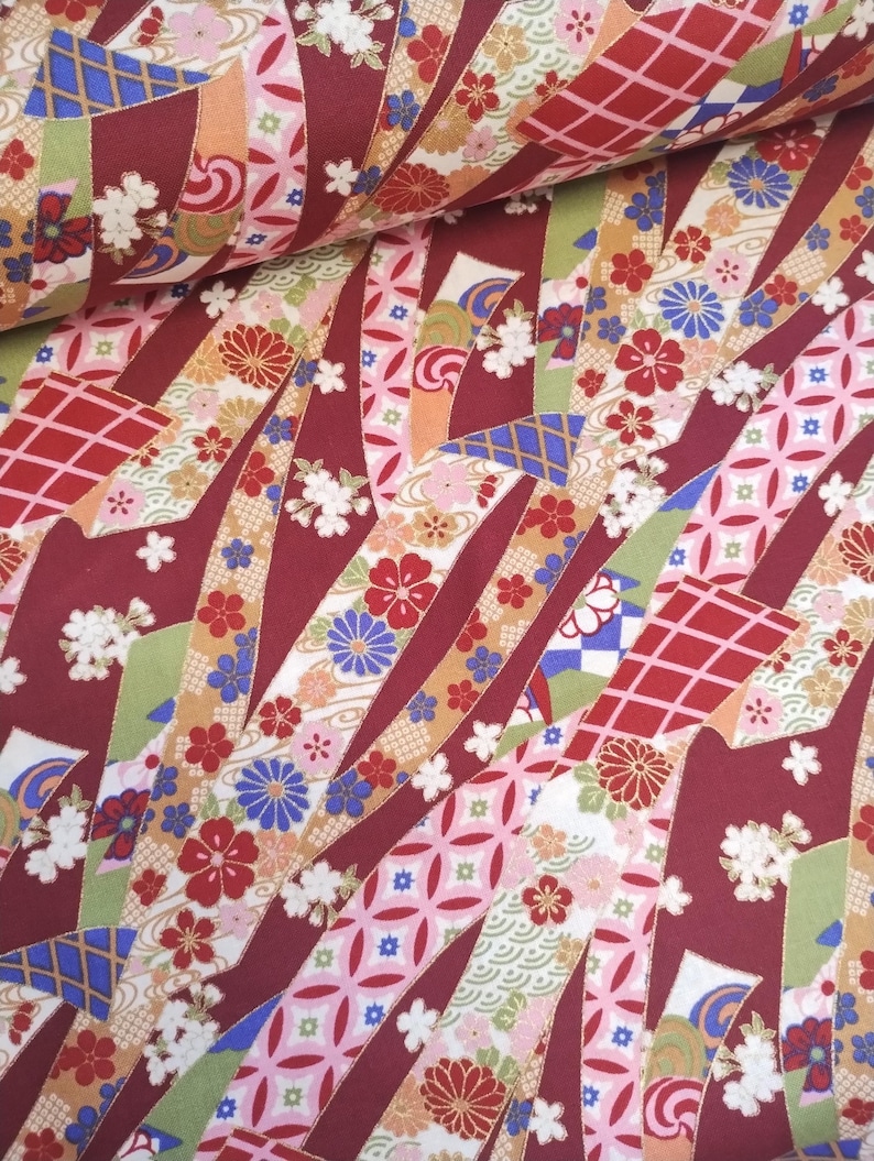 May include: Red fabric with a repeating pattern of colourful ribbons and flowers. The ribbons are arranged in a diagonal pattern and are decorated with gold accents. The flowers are in shades of pink, blue, and white.