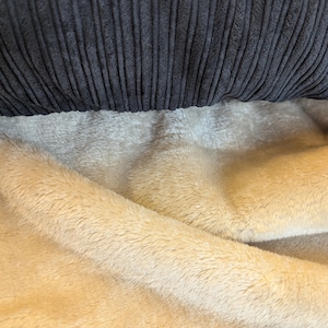 Könnte beinhalten: Nahaufnahme eines dunkelgrauen Cordstoffs über einem beigen, flauschigen Material. Der Cord hat vertikale Rippen, und der beige Stoff hat eine weiche, plüschige Textur. Der Kontrast in Texturen und Farben erzeugt ein gemütliches Gefühl.