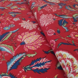 Op de afbeelding: Rode stof met een gedetailleerd bloemenpatroon. Het ontwerp toont grote roze en witte bloemen, blauwe en groene bladeren en kleinere gele en blauwe bloesems. De stof is gevouwen en toont de herhaling van het patroon.