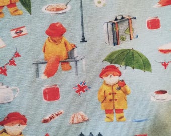May include: A blue fabric with a repeating pattern of red foxes wearing yellow raincoats and holding umbrellas. The pattern also includes red and white flags, streetlights, suitcases, and other London-themed imagery.