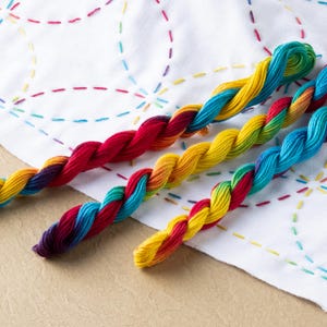 Op de afbeelding: Drie bollen regenboogkleurige borduurgaren op een witte stoffen achtergrond. Het garen is gedraaid en heeft een kleurverloop van rood naar geel naar blauw.
