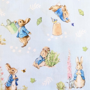 May include: Light blue fabric featuring a repeating pattern of brown rabbits wearing blue jackets. The rabbits are depicted in various garden activities, including holding a green watering can, jumping, and standing near pink flowers.