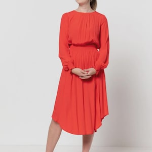 May include: A vibrant red dress with long sleeves and a gathered waist. The dress features a flowing, asymmetrical hemline and is paired with neutral-toned heels. The model is standing in front of a white background.