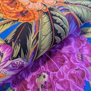 May include: Close-up of a fabric with a vibrant floral pattern. The design features large orange and purple flowers with green leaves against a bright blue background. The fabric is folded, showing the detailed print.