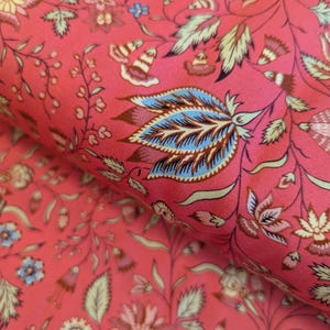 Op de afbeelding: Close-up van een koraalkleurige stof met een gedetailleerd bloemenpatroon. Het ontwerp bevat verschillende bloemen, bladeren en stelen in tinten blauw, groen en crème. De stof heeft een zachte, getextureerde uitstraling, geschikt voor naaiprojecten.
