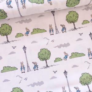 May include: White fabric featuring a repeating pattern of rabbits in blue jackets, trees, street lamps, and clouds. The illustrations are in a whimsical style, with soft green and gray accents. The overall design evokes a storybook or children's theme.
