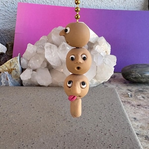 May include: A whimsical, handmade clay sculpture featuring three stacked, expressive faces. The top face has a prominent nose, the middle face has a surprised expression, and the bottom face has a tongue sticking out. The sculpture hangs from a gold chain.