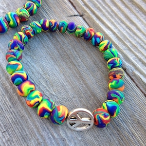 May include: A colorful beaded bracelet with a peace sign charm. The beads are made of a marbled material with shades of blue, green, yellow, and red.
