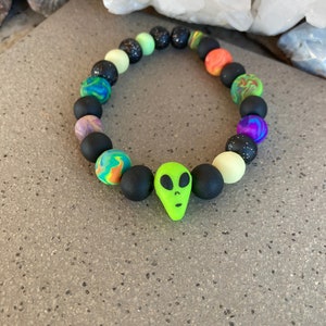 May include: A colorful beaded bracelet with a green alien charm. The beads are black, green, orange, purple, and blue. The bracelet is made of polymer clay.