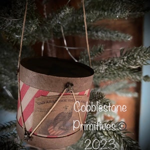 May include: A small, brown, paper-wrapped drum with red and white stripes. The drum has a vintage image of an eagle and the text "Old Abe the War Eagle". The drum is hanging from a string and is decorated with small metal stars. The text "Cobblestone Primitives © 2023" is visible in the image.