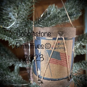 May include: A small, brown, fabric drum with a blue and white American flag design. The drum is hanging from a string and has the text "Cobblestone Primitives © 2023" on it.