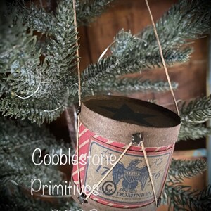 May include: A small, brown paper drum with red and white stripes. The drum has a star on the top and a black and white image of an eagle with the text "1776" and "1770" on either side. The drum is hanging from a string and is decorated with small metal stars.