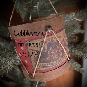 May include: A small, brown, paper-wrapped drum ornament with a red, white, and blue design. The drum has a red, white, and blue flag with stars and the text "1776 Bicentennial" on it. The drum is hanging from a string on a Christmas tree.