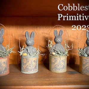 May include: Four gray bunny figurines with wire handles are displayed on a wooden shelf. Each bunny is holding a small basket filled with straw and a few small eggs. The baskets are decorated with a vintage-style print of bunnies and eggs. The text "Cobblestone Primitives© 2023" is visible at the top of the image.