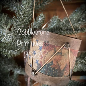 May include: A small, brown, fabric-covered drum with a vintage American flag design. The drum is decorated with silver stars and is hanging from a string on a green Christmas tree.