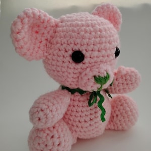 May include: A handmade pink crochet teddy bear with black button eyes and a green ribbon around its neck. The bear has a textured, stitched appearance and is sitting upright. The toy is a soft, plush item.