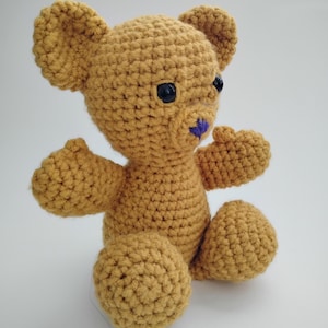 May include: A golden-brown crochet teddy bear with black button eyes and a purple nose. The handmade bear has rounded arms and legs, and is sitting upright. A charming, soft toy, perfect for a child.