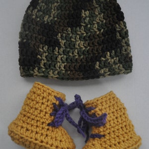 May include: A crochet beanie featuring a camouflage pattern in shades of dark green, brown, and black, paired with a set of mustard yellow crochet booties that include purple yarn laces tied in a bow at the front of each shoe.