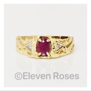 Puede incluir: Un anillo de oro con un rubí rojo y dos pequeños diamantes. El anillo tiene un diseño texturizado.