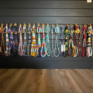 May include: A wall display of colourful beaded necklaces, each with a unique pattern and colour combination. The necklaces are hung on hooks against a dark wood background.