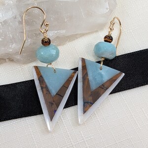 May include: A pair of handcrafted earrings featuring gold-colored ear wires, light blue gemstone beads, and triangular pendants. The pendants have a light blue center, a brown wood-grain design, and white edges. The earrings are displayed on a white surface.