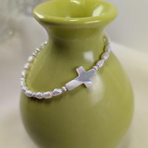 May include: A white pearl bracelet with a white mother of pearl cross charm. The bracelet is on a green vase.