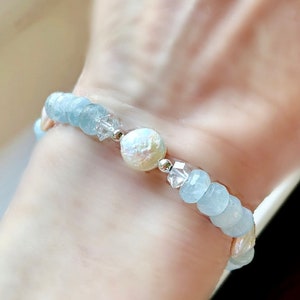 May include: A delicate bracelet featuring light blue aquamarine beads, a white pearl, and clear crystal accents. The bracelet is made with silver spacers and is shown on a person's wrist.