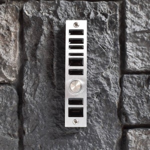 May include: A silver doorbell with a round button and eight rectangular buttons. The doorbell is mounted on a stone wall.