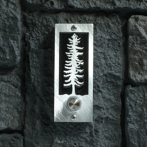 May include: A rectangular doorbell button with a brushed metal finish. The button features a black inset with a white tree silhouette. A round, silver button is centered below the tree design, and the doorbell is mounted on a dark gray stone wall.