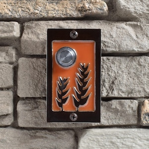 May include: A black metal doorbell button with an orange background and a stylised design of two plants. The button has a round, silver-coloured button in the centre.