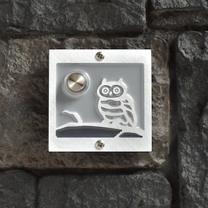 May include: A white square doorbell with a silver button and a gray owl design. The owl is perched on a branch with a moon in the background.