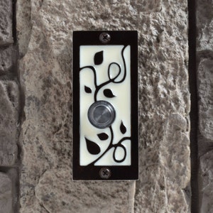 May include: A white ceramic doorbell button with a black floral design and a round, silver button. The doorbell is mounted on a stone wall.