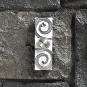 May include: A silver doorbell with a decorative swirl design and a round button. The doorbell is mounted on a stone wall.