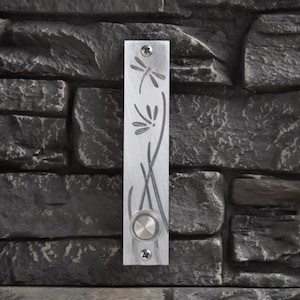 May include: A silver doorbell button with a dragonfly and floral design. The button is mounted on a brick wall.